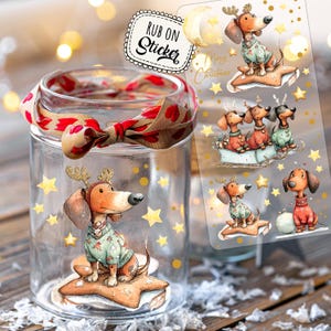 Op de afbeelding: Een heldere glazen pot met een rood en bruin lint om de bovenkant. In de pot zit een sticker van een teckel met een trui en geweien, zittend op een ster. Een vel stickers met teckels in kerstoutfits is zichtbaar.