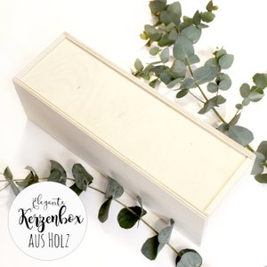May include: A white wooden box with the text "elegante Kerzenbox AUS HOLZ" on a white circle sticker. The box is surrounded by green eucalyptus branches.