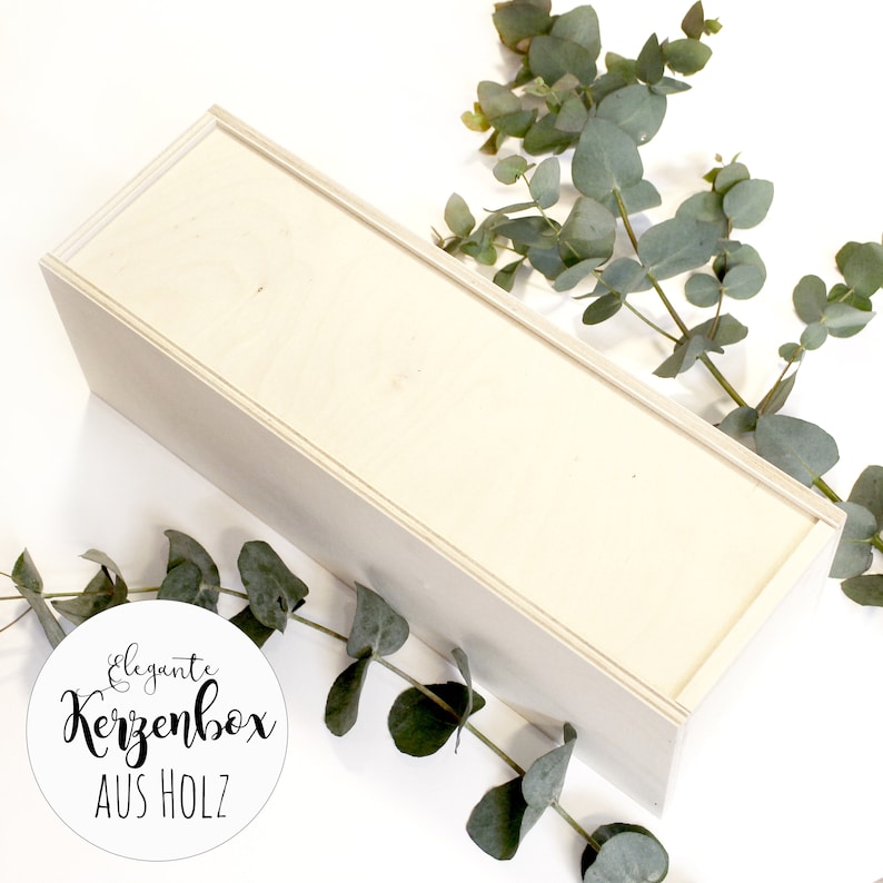 May include: A white wooden box with the text "elegante Kerzenbox AUS HOLZ" on a white circle sticker. The box is surrounded by green eucalyptus branches.