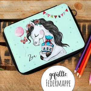 Op de afbeelding: Een mintgroene etui met een witte paardenillustratie die een cadeaudoos vasthoudt met de letters ABC erop. Het paard draagt een roze bloemenkroon en een roze ballon zweeft erboven. De woorden "Zoe" en "gefüllte FEDERMAPPE" staan op het etui gedrukt.