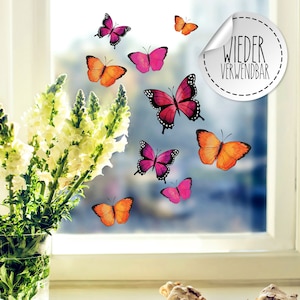 Op de afbeelding: Een set herbruikbare vlinder-raamstickers in roze, oranje en paars. De stickers zitten op een raam met een boeket witte bloemen in een glazen vaas. De tekst "WIEDER VERWENDBAR" staat op een witte ronde sticker.