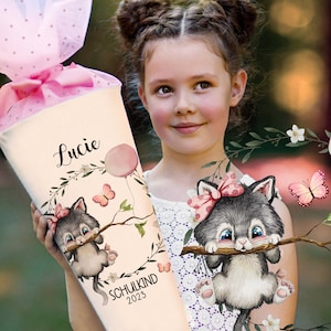 Könnte beinhalten: Ein weißer Schultüte mit einer rosa Schleife mit weißen Punkten. Die Tüte hat einen rosa Ballon, Schmetterlinge und ein graues Kätzchen mit einer rosa Schleife, das an einem Ast hängt. Die Tüte ist mit dem Namen "Lucie" und dem Text "Schulkind 2023" personalisiert.