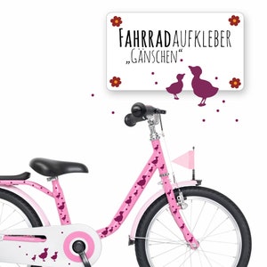 以下が含まれることがあります： 白いフレームと紫色のアヒルステッカーが付いたピンクの子供用自転車。自転車には黒いサドル、黒いハンドルバー、ハンドルバーに取り付けられた白い旗があります。自転車は「Fahrradaufkleber "Gänschen""と書かれた看板で飾られています。