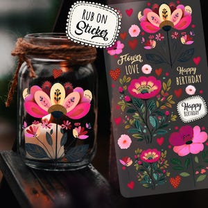 Könnte beinhalten: Ein Glas mit einem Blumendesign und einer brennenden Kerze im Inneren. Das Glas ist mit einem rosa und gelben Blumenaufkleber verziert. Ein Bogen mit Rubbelbildern mit Blumendesigns und den Worten "Flower Love" und "Happy Birthday" befindet sich neben dem Glas.