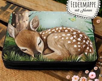 Könnte beinhalten: Ein rechteckiges Federmäppchen mit schwarzem Reißverschluss zeigt eine Illustration eines schlafenden Rehs. Das Reh ist braun mit weißen Flecken und liegt im grünen Gras mit einem Baum und Lichterketten im Hintergrund. Der Text "GEFÜLLTE FEDERMAPPE mit Namen" steht oben rechts.
