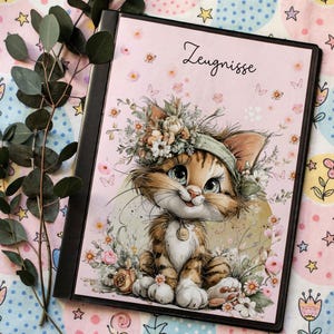 Puede incluir: Una carpeta negra con una cubierta rosa que presenta un gatito de dibujos animados con una corona floral. El gatito está rodeado de flores y mariposas. La palabra "Zeugnisse" está escrita en cursiva en la parte superior.