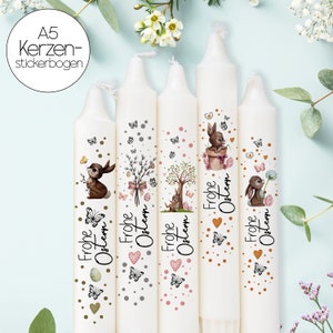 以下が含まれることがあります： 装飾的なデザインの白いイースターキャンドル5本。各キャンドルには、ウサギ、蝶、そして「Frohe Ostern」の言葉の異なるイラストが描かれています。キャンドルはハートとドットで飾られています。