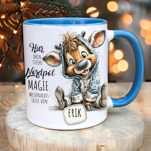 Może przedstawiać: Biały ceramiczny kubek z niebieskim wnętrzem i uchwytem. Kubek przedstawia kreskówkowego renifera w niebieskiej bluzie z kapturem. Tekst na kubku brzmi "Hier drin steckt Nordpol Magie Weihnachts-Tasse von Erik."