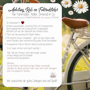 May include: A white bicycle with a brown seat and daisy decals is shown. A white card with German text provides instructions for applying rub-on bicycle stickers. The card includes the text "Anleitung Rub on Fahrradsticker".