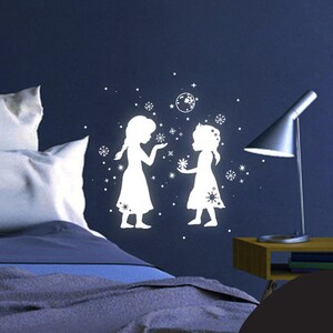 Könnte beinhalten: Weiße Silhouette-Wandaufkleber von zwei Mädchen, die sich an den Händen halten, mit Schneeflocken um sie herum. Der Aufkleber befindet sich an einer dunkelblauen Wand mit einem weißen Bett und einer Lampe im Hintergrund.