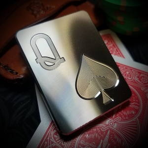 May include: A silver metal Queen of Spades playing card charm with a detailed spade design and a stylized Queen symbol.