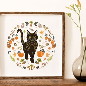 Black Cat Art Print shown in a dark wood frame.. Halloween Folk Art style Autumn Cat Wall Art. Scandinavian style illustration of a black cat surrounded by pumpkins, Chinese lanterns, leaves and berries, in warm autumn colours.