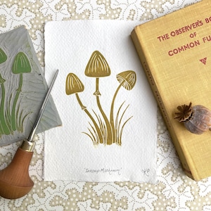 May include: A gold ink stamp print of three mushrooms with stems and leaves on a white background. The print is titled "Inkcap Mushroom".