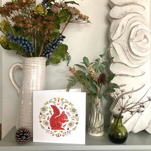 Red Squirrel Card shown on a mantel shelf with jug of dried seedheads and berries.