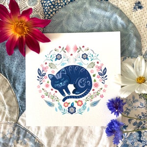 Colourful Cat Greeting Card - Nordic folk art style. It features a sleeping cat surrounded by flowers, ferns and leaves in  blues, pinks and turquoise. Photographed on a blue and cream patchwork quilt.