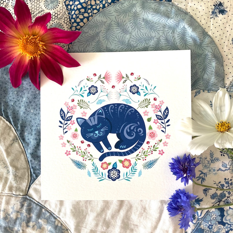 Colourful Cat Greeting Card - Nordic folk art style. It features a sleeping cat surrounded by flowers, ferns and leaves in  blues, pinks and turquoise. Photographed on a blue and cream patchwork quilt.