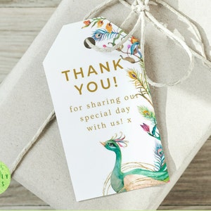 Puede incluir: Una etiqueta de regalo blanca con texto dorado que dice "¡Gracias! por compartir nuestro día especial con nosotros! x". La etiqueta está decorada con una ilustración de acuarela de un pavo real con plumas verdes, azules y doradas. La etiqueta está atada con un cordel a una bolsa de regalo de papel marrón con una pegatina circular verde que dice "IMPRESIONES ECOLÓGICAS".