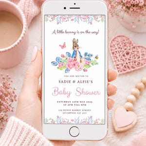 May include: A digital baby shower invitation displayed on a smartphone screen featuring a watercolor illustration of a bunny in a blue jacket, pink flowers, a butterfly, and a bee. The text reads A little bunny is on the way! Sadie & Alfie's Baby Shower, Saturday 12th May 2025 at 3pm till 8pm, 123 Bunny Rabbit Street, Liverpool L37 4GV.