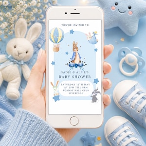 May include: A smartphone displaying a digital baby shower invitation with a blue theme, featuring illustrations of rabbits, a hot air balloon, and the text: YOU'RE INVITED TO SADIE & ALFIE'S BABY SHOWER SATURDAY 12TH MAY AT 3PM TILL 8PM FORMBY HALL CLUB LIVERPOOL.