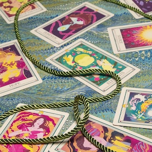 May include: A collection of tarot cards with colorful illustrations and text, arranged on a patterned surface. A green and gold rope is looped around some of the cards. The cards feature images of figures, planets, and abstract designs.