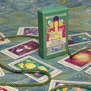 May include: A green box of "The Dark Earth Oracle" cards with illustrations of figures and celestial bodies. The cards are spread out on a blue and green patterned surface, with a green rope-like cord. The box and cards feature colorful artwork.
