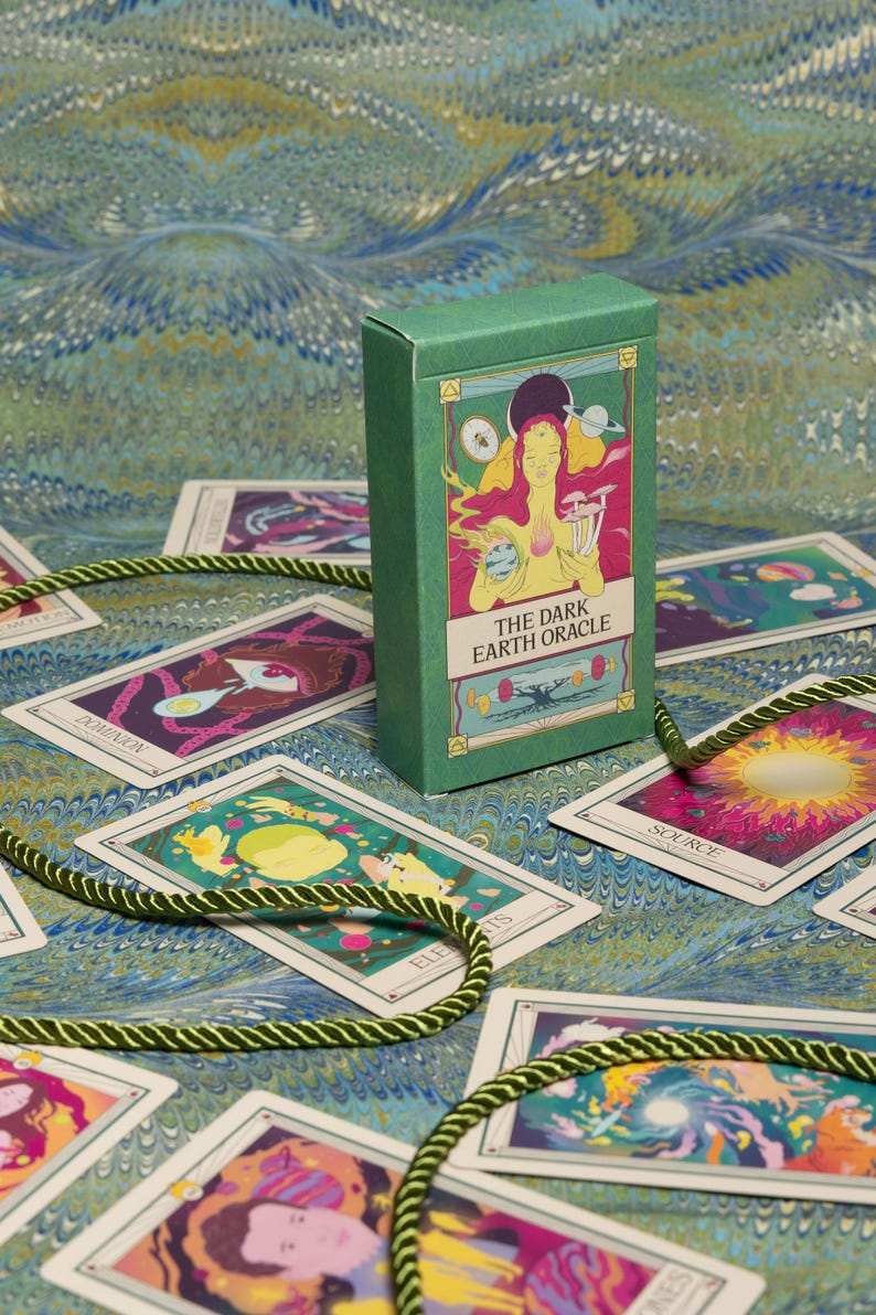 May include: A green box of "The Dark Earth Oracle" cards with illustrations of figures and celestial bodies. The cards are spread out on a blue and green patterned surface, with a green rope-like cord. The box and cards feature colorful artwork.