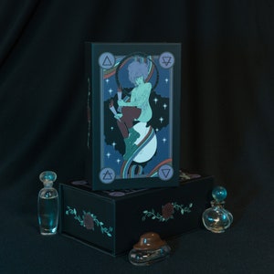 May include: A rectangular black box featuring a graphic illustration of a person with purple hair sitting on a crescent moon, surrounded by stars and elemental symbols. Three small glass perfume bottles with stoppers are placed in front of the box.