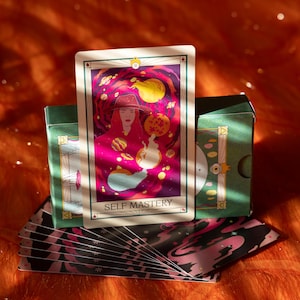 May include: A tarot card with the words "SELF MASTERY" is displayed, featuring a woman in a hat against a cosmic background. The card is part of a deck with a green box and a patterned design. The image has a warm, orange and pink color scheme.
