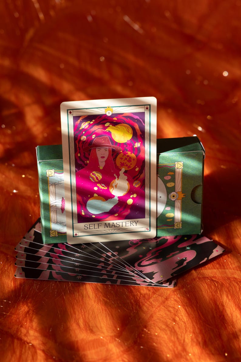 May include: A tarot card with the words "SELF MASTERY" is displayed, featuring a woman in a hat against a cosmic background. The card is part of a deck with a green box and a patterned design. The image has a warm, orange and pink color scheme.