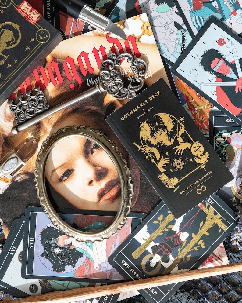 May include: A Gothmancy Deck guide book with a black cover and gold illustration, an ornate silver-toned skeleton key, a small oval metal frame, and several tarot cards featuring graphic illustrations, including The Hanged Man card.