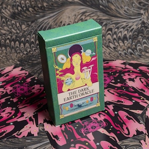 May include: A green box of "The Dark Earth Oracle" cards with a colorful illustration of a woman, planets, and mushrooms. The cards are scattered around the box, featuring a black and pink design with geometric shapes and stars. The background has a marbled pattern.