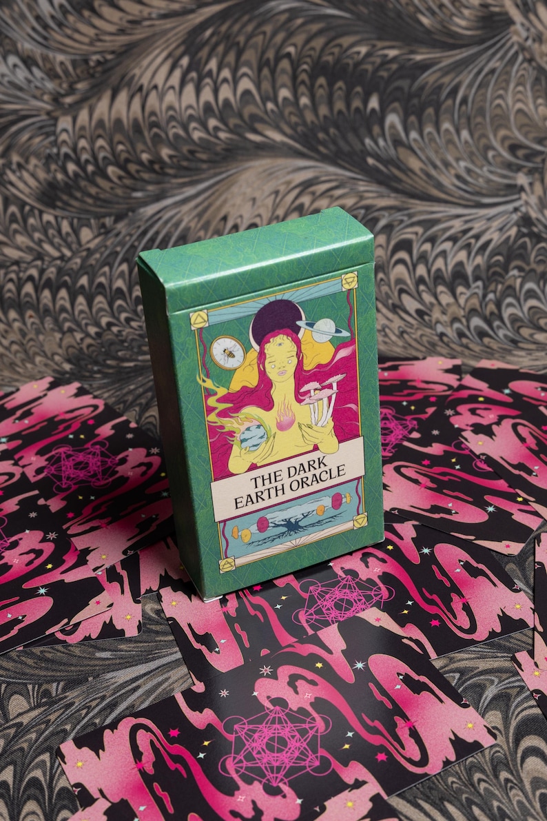 May include: A green box of "The Dark Earth Oracle" cards with a colorful illustration of a woman, planets, and mushrooms. The cards are scattered around the box, featuring a black and pink design with geometric shapes and stars. The background has a marbled pattern.