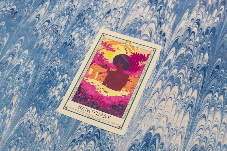 May include: A tarot card with the word "SANCTUARY" at the bottom. The card features a colorful illustration of a person in a tropical setting with pink, orange, and yellow hues. The background is a blue and white marbled pattern.