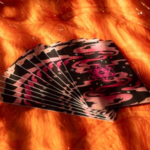 May include: A fan of tarot cards with a black background and pink cloud-like designs. The cards have a pink geometric symbol and white star accents. The cards are fanned out on an orange, textured surface.