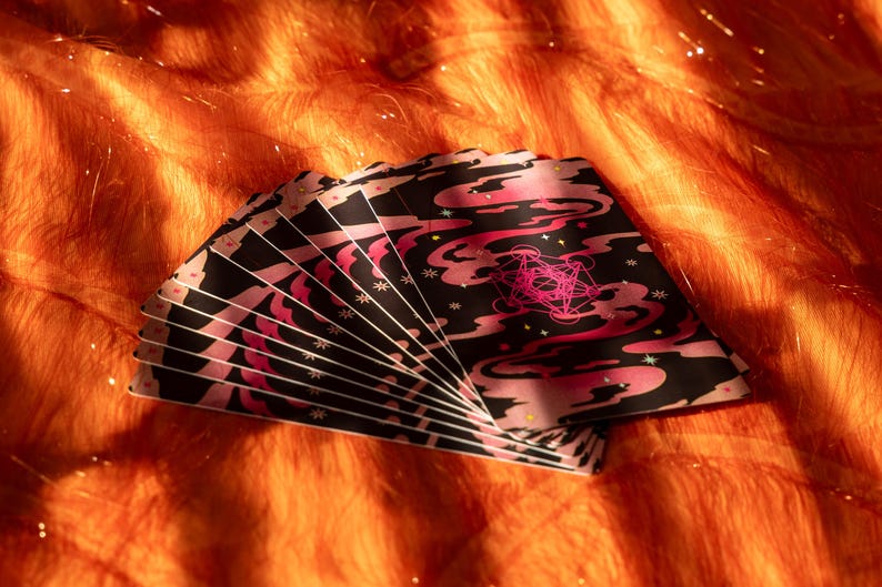 May include: A fan of tarot cards with a black background and pink cloud-like designs. The cards have a pink geometric symbol and white star accents. The cards are fanned out on an orange, textured surface.