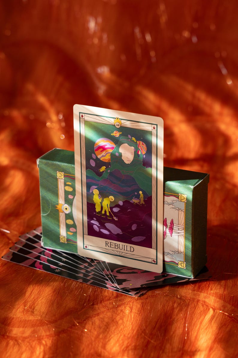 May include: A tarot card with the word "REBUILD" and a green box. The card features an illustration of a person with a green cloak and a planet. The box is green with gold accents. The background is orange.