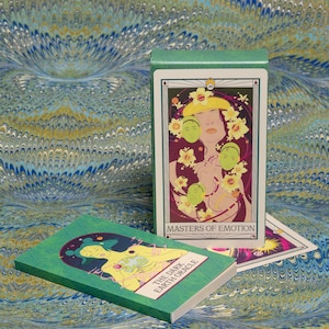 May include: A green tarot card box and deck with the text "Masters of Emotion" and "The Dark Earth Oracle". The cards feature illustrations of figures with floral and celestial elements. The background is a marbled pattern in shades of blue, green, and gold.