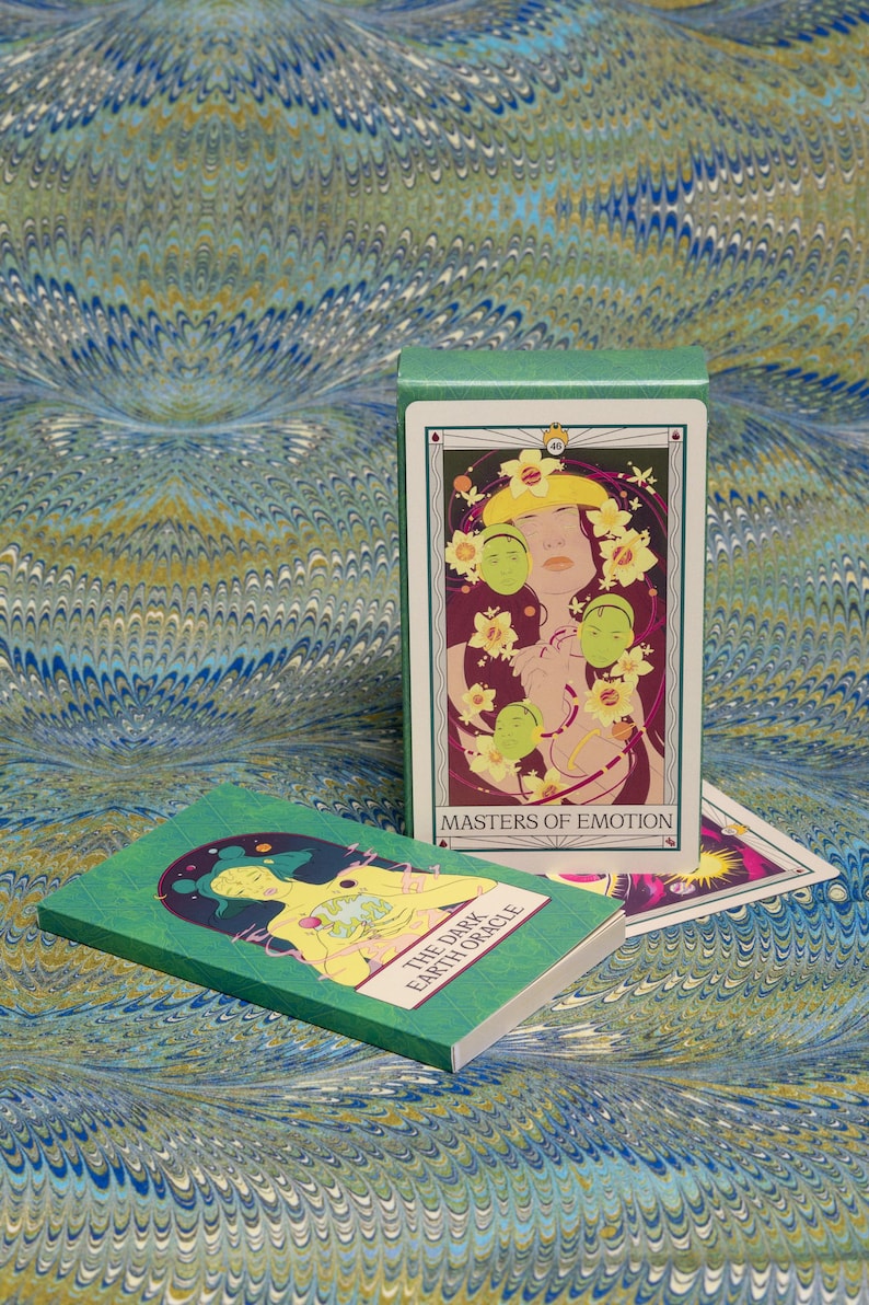May include: A green tarot card box and deck with the text "Masters of Emotion" and "The Dark Earth Oracle". The cards feature illustrations of figures with floral and celestial elements. The background is a marbled pattern in shades of blue, green, and gold.