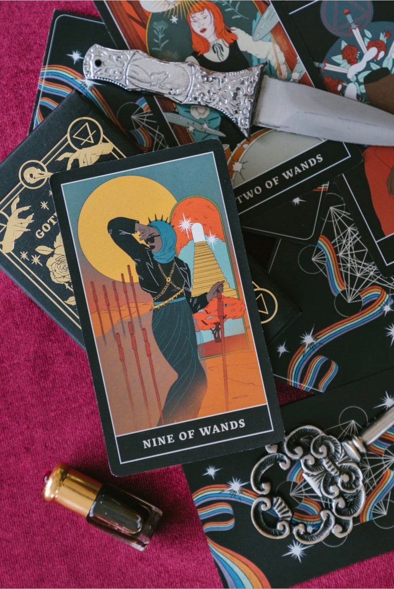 May include: A Nine of Wands tarot card featuring a figure in a black outfit and blue head covering, surrounded by eight wands, with a large yellow sun and a staircase in the background. A silver dagger and an ornate silver key rest on the cards.