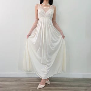 May include: A floor-length cream-colored gown featuring a sheer mesh neckline, a sweetheart bodice with intricate lace detailing, and a flowing skirt. The garment is paired with sheer, elbow-length gloves and matching lace-covered pointed-toe heels.