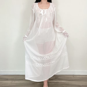 以下が含まれることがあります： 長い袖とレースのトリムが付いた白いシアーなマキシドレス。ドレスはギャザーネックラインとフロントにタイが付いています。