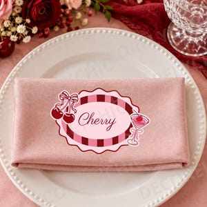 May include: A folded dusty pink cloth napkin centered on a white plate, featuring a decorative oval label with a dark pink and red checkered border, the word Cherry in script, a pair of cherries with a bow, and a cocktail glass with a cherry.