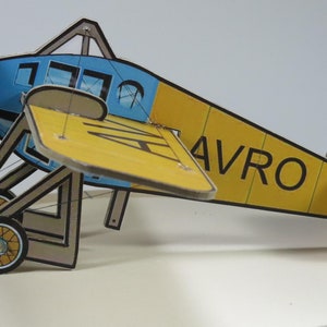 以下が含まれることがあります： 側面に「AVRO」と印刷された黄色と青色の段ボール製飛行機の模型。模型にはプロペラ、翼、およびランディングギアがあります。