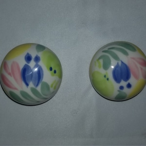 May include: Two round ceramic knobs with a white background and colourful floral designs. The designs feature pink, yellow, green, and blue colours.