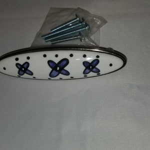 Könnte beinhalten: Weißer Keramik-Schrankknauf mit silberner Einfassung und drei lila Blüten mit schwarzen Umrissen. Der Knauf wird mit vier silbernen Schrauben geliefert.