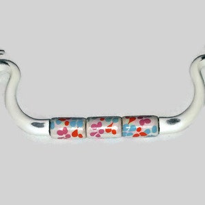 Könnte beinhalten: Weißer Metall-Schubladenzug mit drei bunten Keramikknöpfen. Die Knöpfe haben ein Blumenmuster in Rosa, Rot und Blau.