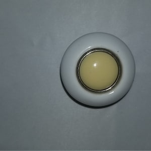 Pode incluir: Um botão redondo branco com um centro amarelo, montado em uma superfície cinza claro.