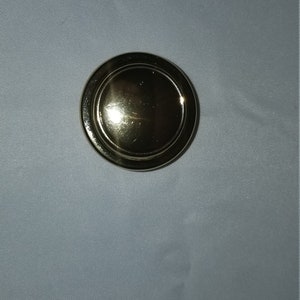 May include: A gold-colored round knob with a raised center and a smooth surface.