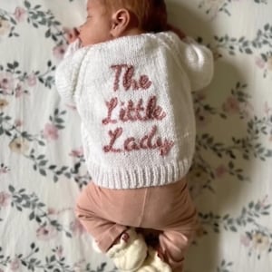 Puede incluir: Un bebé recién nacido con un suéter de punto blanco con el texto bordado "The Little Lady" en rosa. El bebé también lleva pantalones rosas y botines blancos, acostado sobre una manta con estampado floral.