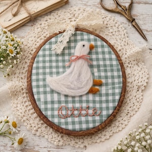 May include: A circular wall hanging with a white goose design, orange beak and feet, and a pink bow, set against a green and white gingham pattern. The name "Ottie" is embroidered below. The wooden frame is decorated with lace and a lace ribbon.